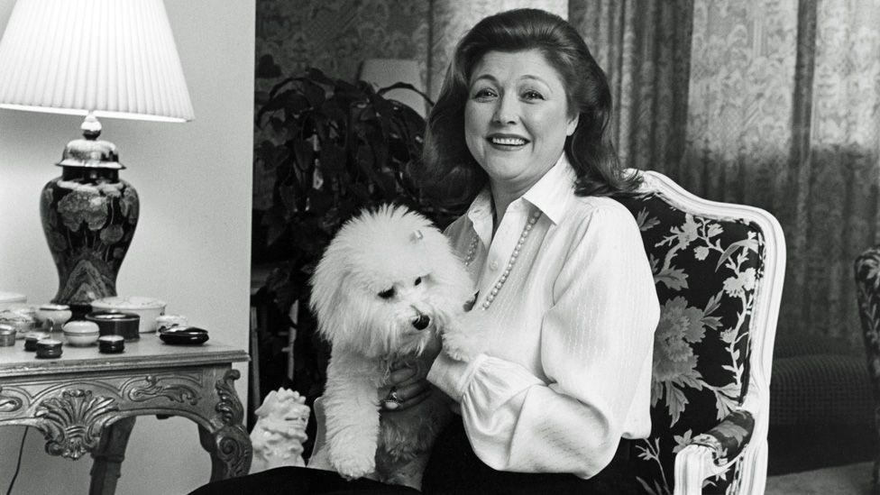 Black-and-white photo of Barbara Taylor Bradford sitting in an ornate chair, smiling with a white poodle on her lap