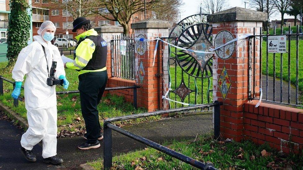 A forensic examiner dressed in a full white suit, blue latex gloves, a mask and a hairnet. They are carrying a camera around their neck and are walking into Rawnsley Park. There is also a female police officer standing near the gate, where blue police tape has blocked off the entrance.