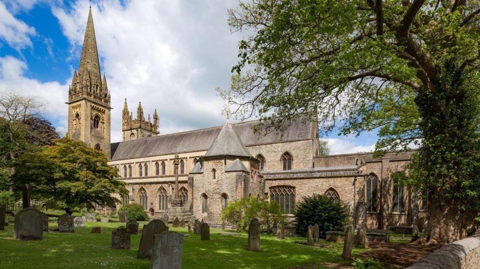 An exterior photo of Llandaff Cathedral. The cathedral is in the background with a graveyard, with headstones in the foreground. There are trees to the left and right of the cathedral.