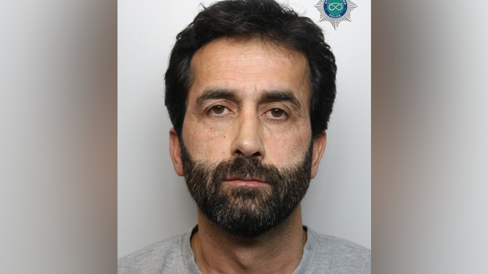 A police mugshot showing a man with a dark beard and short black hair.