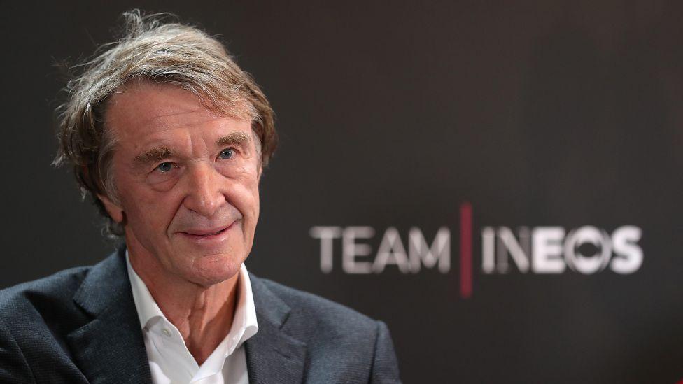 Sir Jim Ratcliffe, with dark greying hair and wearing a dark suit, in a medium close-up shot in front of a "Team Ineos" sign. 