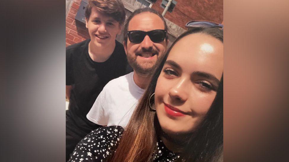 A selfie of Mia with two friends. One has a beard and is wearing dark sunglasses and a white shirt, the other is wearing a dark t-shirt.