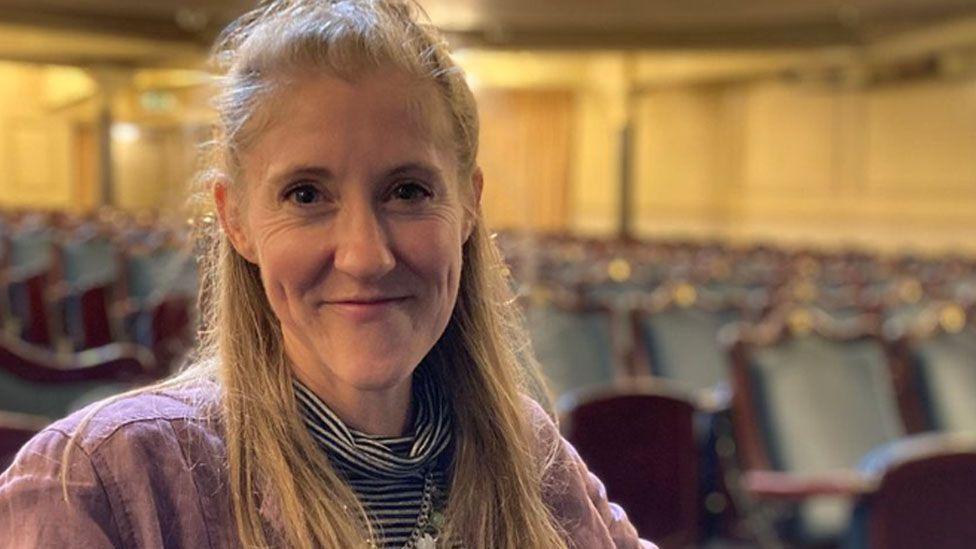 Head-and-shoulders shot of Kerry Vasiliou, sitting in a theatre flooded with  light. She is smiling and has long blonde hair tied back off her face.