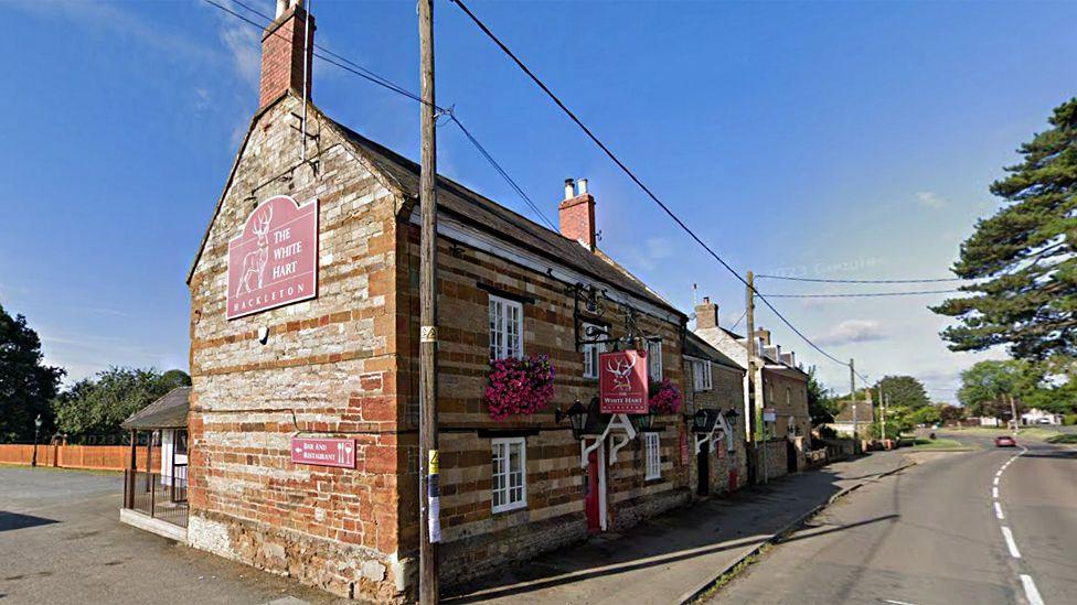 The exterior of the White Hart pub, which is a stone-built two-storey building with small porch over the door. It has big red signs on the side and above the door advertising the pub. There are two big hanging baskets with pink flowers underneath the upper floor windows. It is next to a road and there is a car park behind.