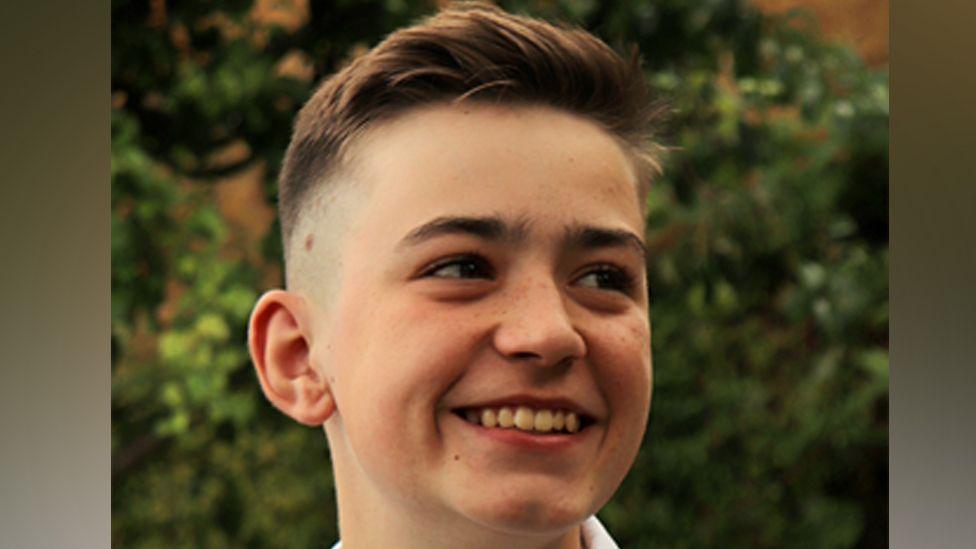 Michal Wojcik, smiling, looking away from the camera, with short dark hair, standing in front of greenery. 
