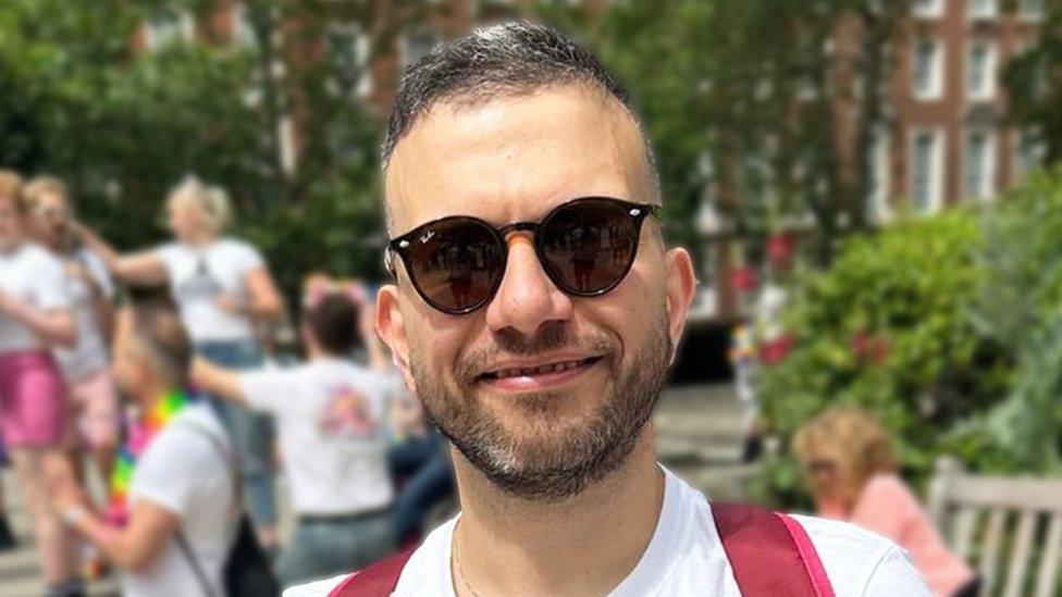 Giancarlo wearing sunglasses and smiling at the camera. He is wearing a white t-shirt and red backpack. The background is blurred, with trees and green hedges visible, along with multiple people.