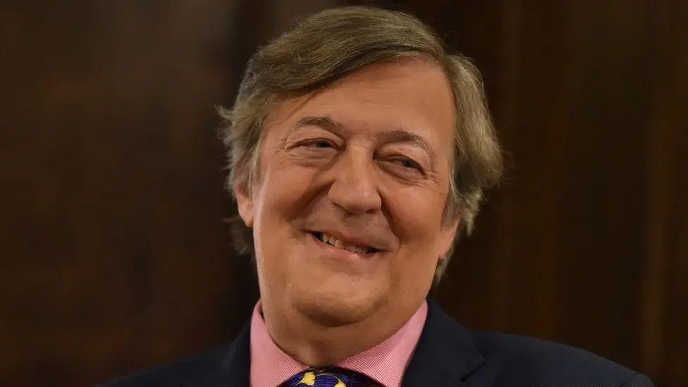 Stephen Fry, with long brown/grey hair, smiles for the camera.