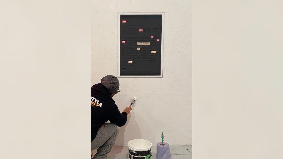 A man in a black hoodie and a grey hat, crouched down and painting a white wall. Above him is one of the pieces of found poetry, framed and with certain words in the text highlighted.