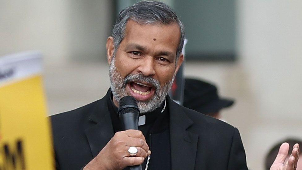 The Right Reverend Doctor John Perumbalath, with a grey beard and dark grey hair, smiles for the camera with trees in the background