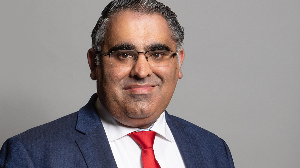 Head and shoulders shot of MP Tahir Ali. He is wearing half-rimmed glasses, a blue suit and a red tie