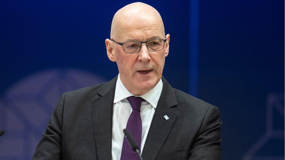 John Swinney, a bald man with glasses wearing a dark suit, white shirt and purple tie, speaks in front of a microphone in a medium close-up. He is standing in front of a blue background. 