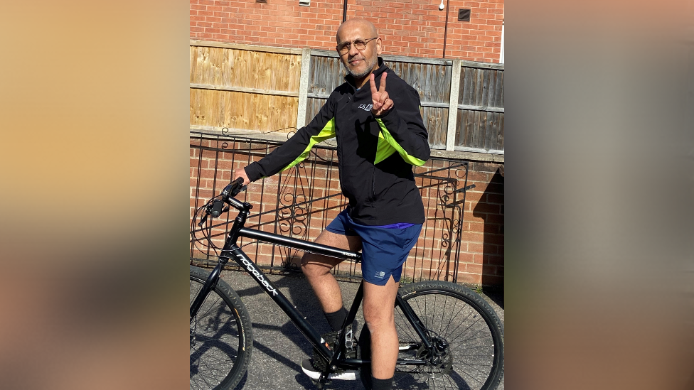 Shahid Akhtar in cycling gear on his bike wearing glasses and making a peace sign with his fingers