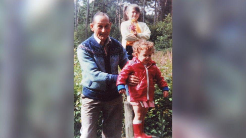 Katrice, in a red coat, boots and skirt, is being held by her grandfather. He has short grey hair and is wearing beige trousers, a white shirt and a blue knitted cardigan. Her older sister Natasha is behind her. She has straight dark hair put up in a ponytail and wears a yellow jumper and white cardigan.