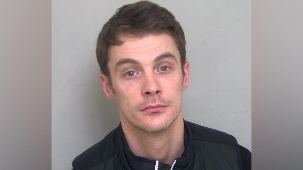 Charlie Martinson is looking at the camera in a police mug shot. He has short, slightly wavy brown hair, a little stubble and is wearing a dark top