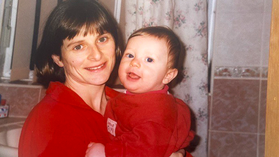 A woman is holding her baby son in her arms. Both mother and son are dressed in red and smiling at the camera. The woman's hair is brown and cut in a bob, with a fringe, and pink bathroom tiles and a floral shower curtain can be seen behind.