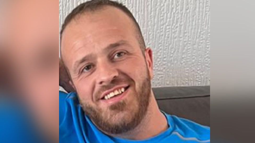 A picture of Craig Richardson smiling looking at the camera. He is wearing a blue t shirt and sat on a sofa. 