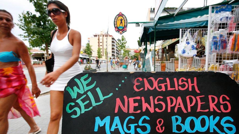 women in beach clothes walking past a sign that reads "We sell English newspapers mags and books"