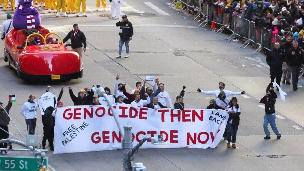 A group of pro-Palestinian protesters stand behind a sign which shows the slogans "genocide then, genocide now" and "free Palestine"