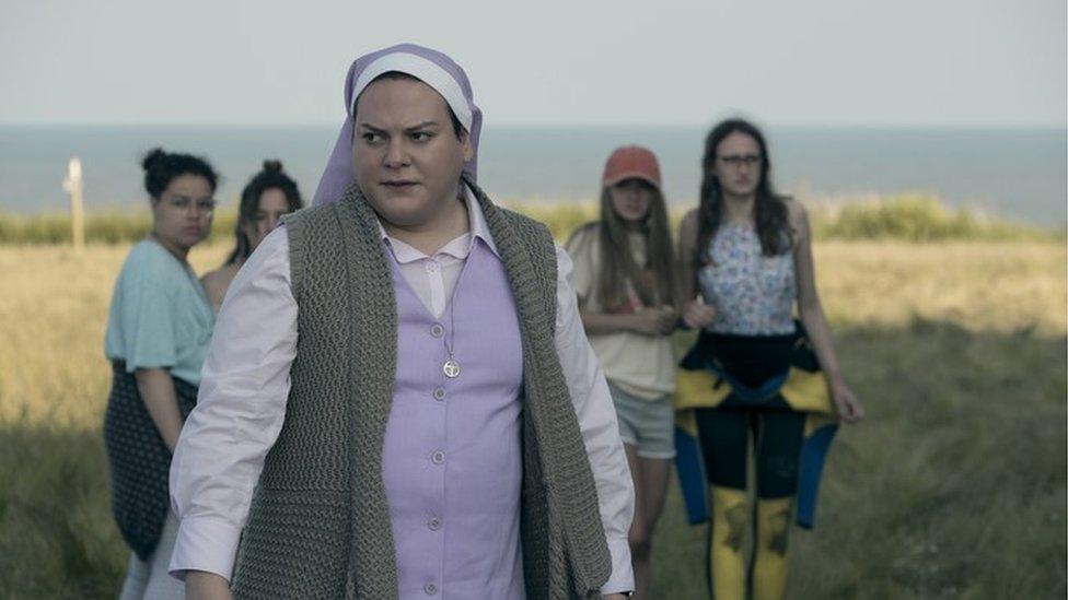 Daniela Vega as Sister Maria, dressed in a nun's habit. Four young girls stand behind here and are out of focus. They appear to be standing on the grassy cliff at Bawdsey