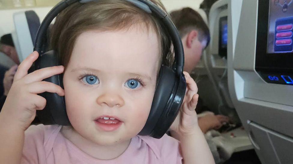 A baby girl, with blue eyes, is photographed while on an aeroplane. She is wearing headphones and is looking directly into the camera.