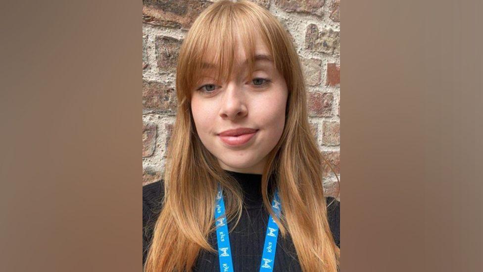 A young woman with blonde-ginger hair and a black top, with a Kyra branded lanyard. In the background is a red brick wall. 