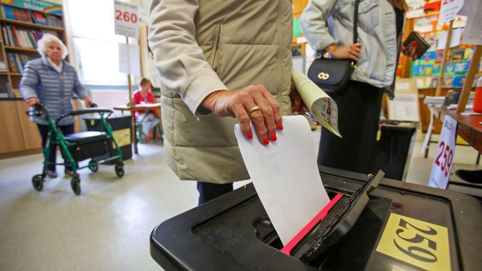 A woman puts a piece of white paper into a black box at a polling station. She is wearing a cream coat and has painted her nails bright pink. The polling station seems to be in a children's library. There is a book case against the wall with lots of colourful books on display. 