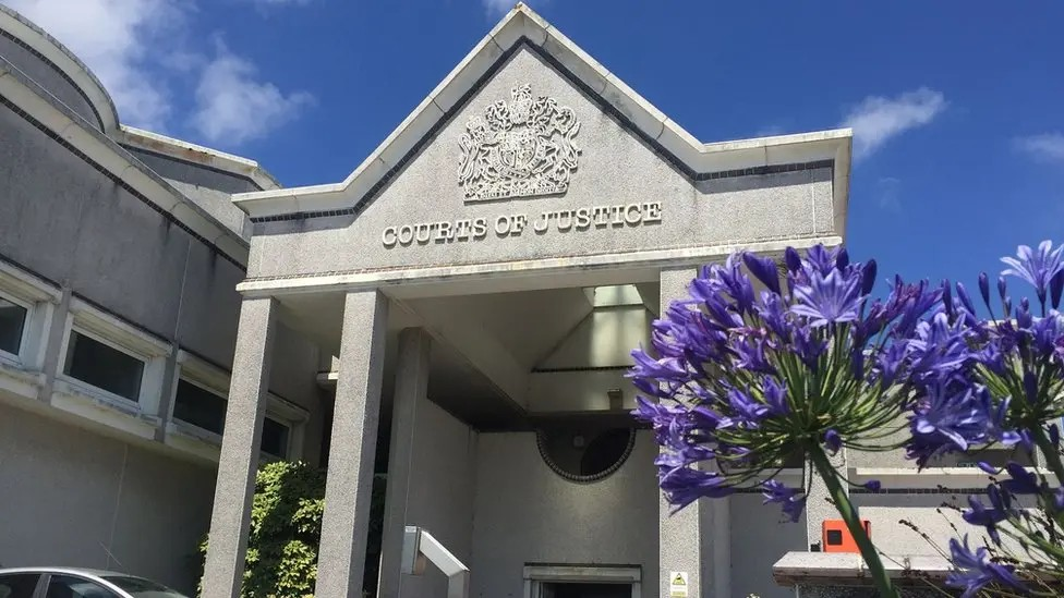 The entrance of a grey court building, with an angular roof and concrete columns. A royal crest and the words Courts of Justice sit above the entrance and purple flowers are in front of the building.