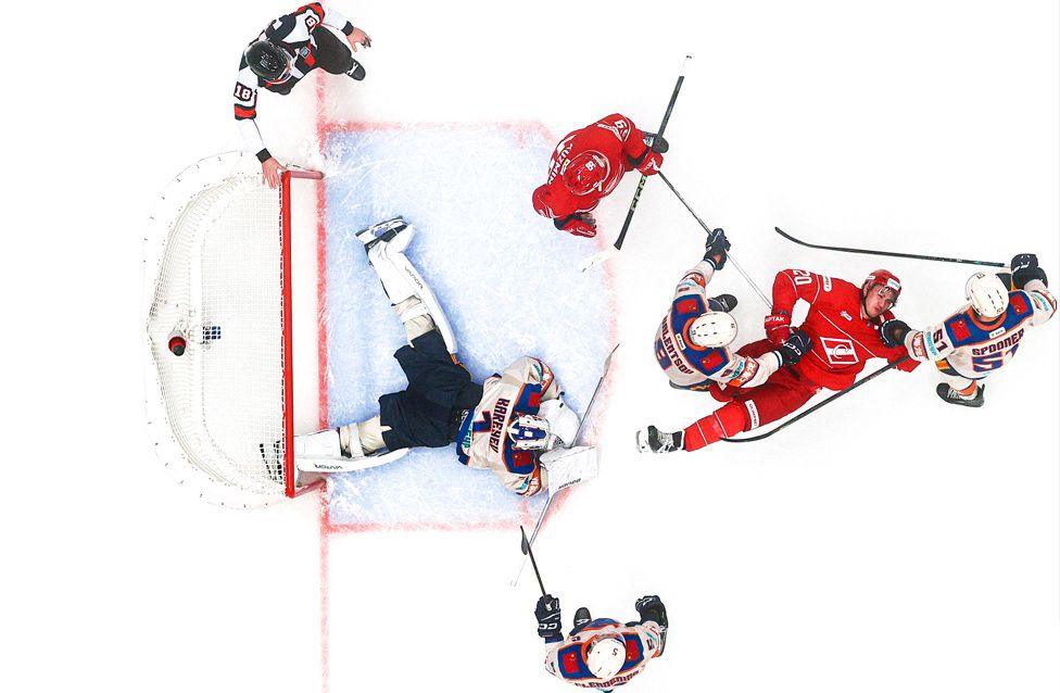 An overhead view of an ice hockey game near the goal. The goalie is sprawled on the ice while players in red and blue jerseys battle for the puck around the crease.