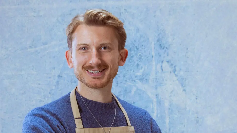 Tom smiling in a dark blue jumper standing against a lighter blue background