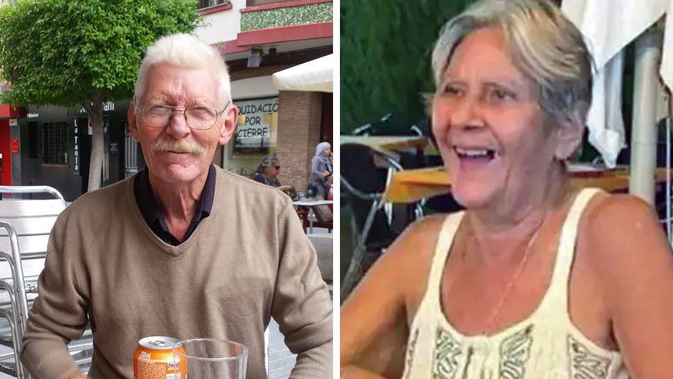 Terry (right) is wearing a white vest top and smiling with a bag on her lap. She has short, grey hair, or her hair is possibly tied back. Don is wearing glasses and has a moustache and is wearing a light brown jumper