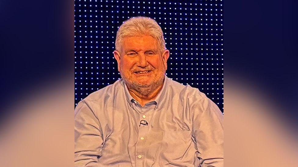 Tim McCarthy has thick white hair and is wearing a light grey open-necked shirt. He is smiling with coloured lights seen behind.