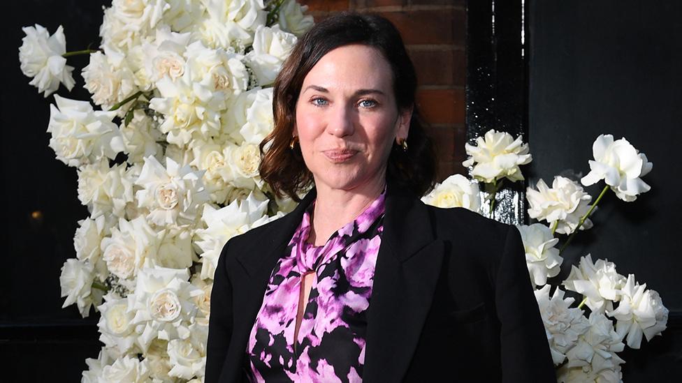 A woman with shoulder length brunette hair stands in front of an elaborate arrangement of white flowers. She's wearing a black jacket over a top with an elaborate, purple-pink floral design