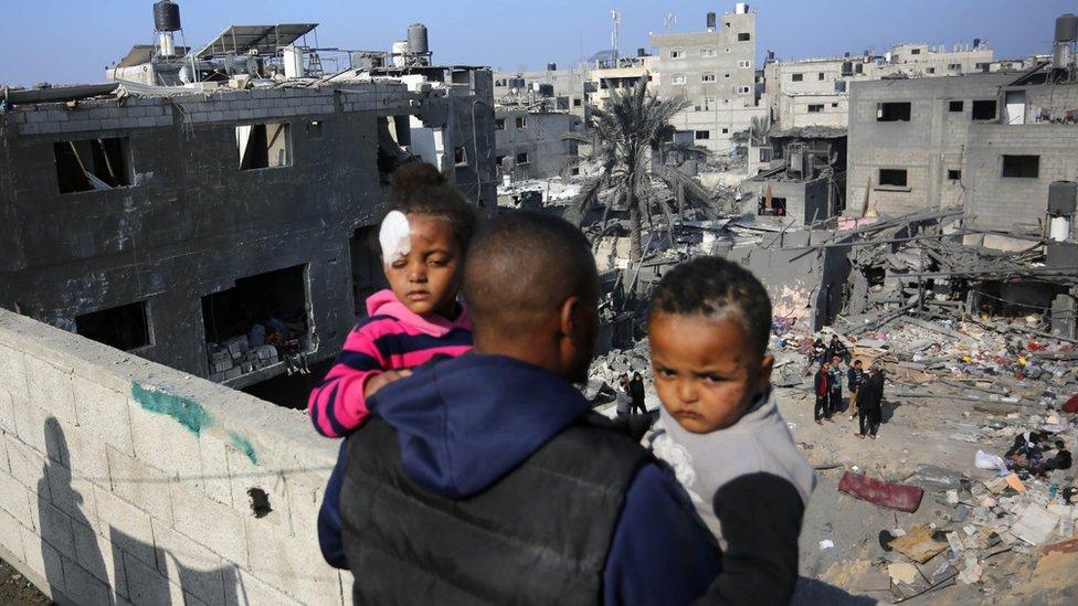 A man holds two children as he looks at the scene of a reported Israeli air strike in Nuseirat refugee camp, in the central Gaza Strip (18 December 2023)