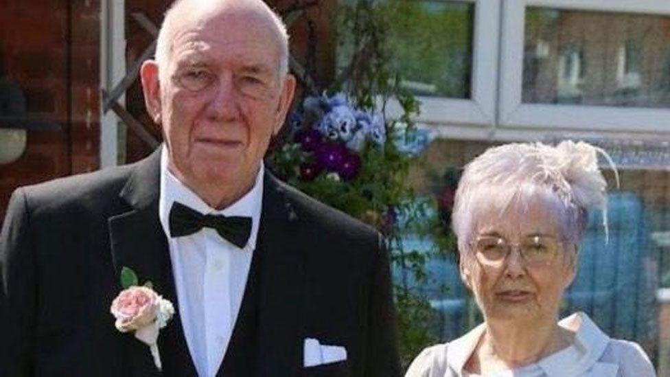 Roy and Susan Pollit pose for a photograph together. Roy wears a formal black suit with a black bow-tie and a pink flower in his button hole. Susan wears a white fascinator and a light grey outfit.