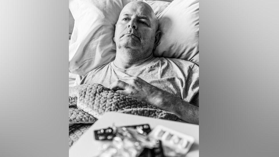 Black and white image of John Newcombe, lying in bed with his hand on his chest, starring into the distance. In front of him are multiple open packets of medication