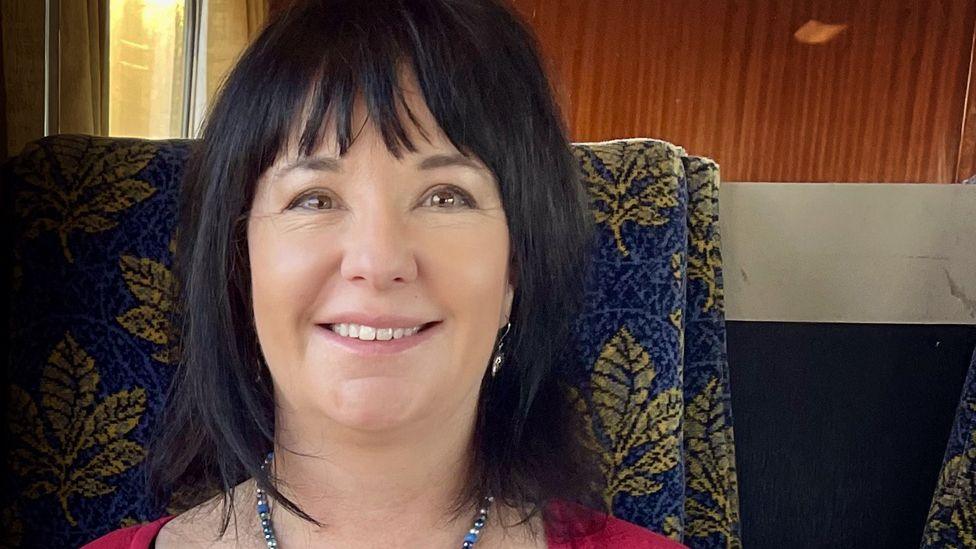 Abi Smith with dark brown hair and a fringe, sitting on a high-backed blue chair with a leaf print on it