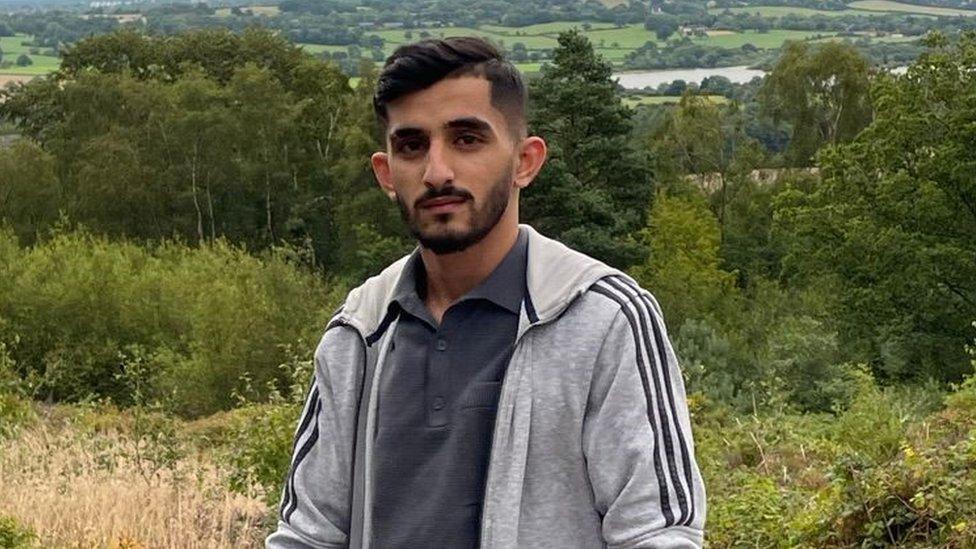 Rizwan Baig pictured on a rural hill surrounded by greenery on an overcast day. Rizwan is an Asian man in his 20s and has short dark hair and a cropped beard. He is wearing a light grey Adidas tracksuit jacket over a dark grey polo shirt. He is looking at the camera with a slight smile