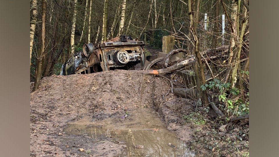 An upturned burned out 4 by 4 vehicle on top of a muddy track, surrounded by woodland.