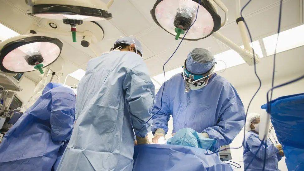 Surgeons in blue scrubs at operating table. There are wires and lights. Three are at the table and one at the endge of the room. A patient is on the table. 