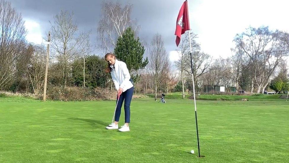 A female golfer makes a putt from about ten feet away from the hole on an outdoor course. She's wearing a white track top and blue leggings, with a look of concentration as the ball rolls towards the black and white pole with a red flag on top inserted into the whole. The well-kept course is surrounded by dozens of trees, most of them missing leaves due to the winter weather.