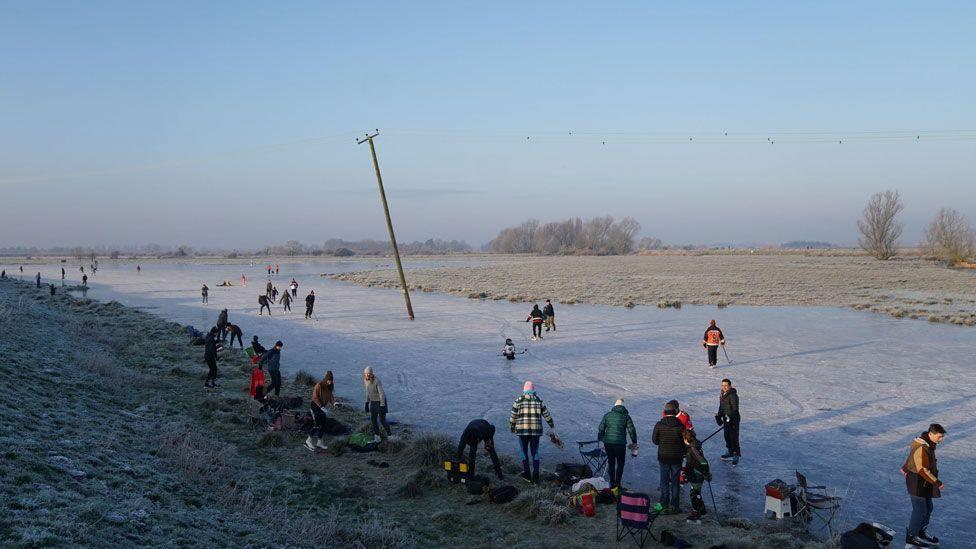 Dozens of people skating on a frozen field at Upware, Cambridgeshire. The blue ice is scored with white lines from the skates. In the middle distance is a field covered in frost and in the far distances are some wintery trees under pale blue skies