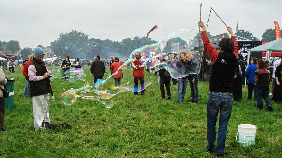 A man with his back to the camera and his arms raised creating a giant soap bubble. He is standing on shaggy grass and is watched by a few people. There are stands and marquees on the right and more marquees, surrounded by people in the distance.