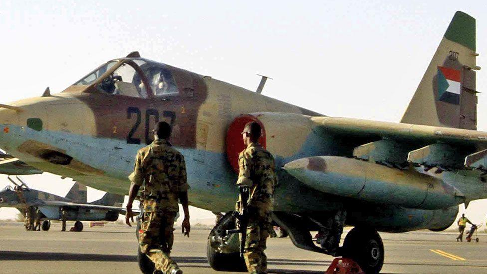 Two men in military uniform walk with their backs to the camera towards a Sudanese war plane on the tarmac at an air base in Sudan in April 2017