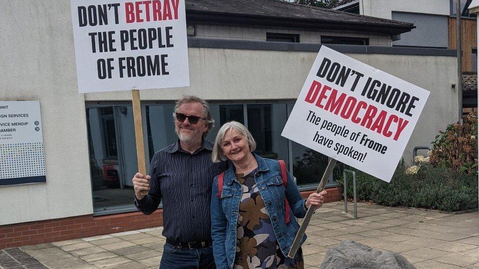 a man and woman holding placards saying 'don't betray the people of Frome' outside a municipal building