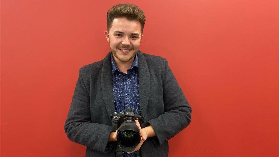 Josh Dury holding a camera and smiling with a red wall behind him