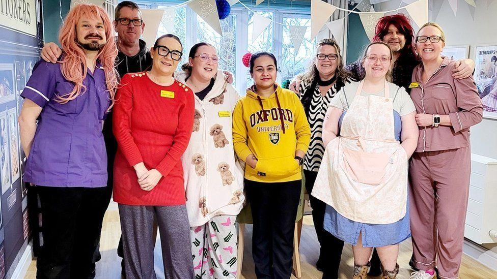 Group of nine adults standing in a line with bunting over them - they are wearing a range of outfits - pyjamas and two are wearing long wigs.