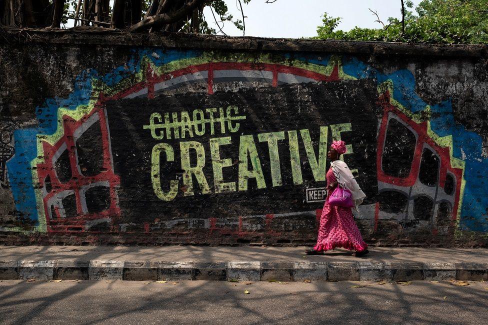 A woman walks near a wall mural that reads 'creative'. The word 'chaotic' is written above it but has been crossed out.