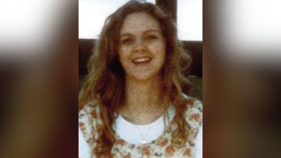 A woman with long, curly brown hair, smiling, she is wearing a white top with a floral pattern