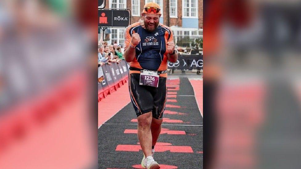 A man celebrates after crossing the finish line at a running event, with people behind a black and red barrier smiling and cheering