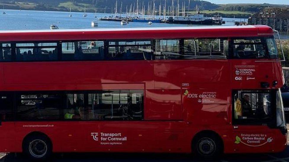 A red double decker electric bus from Go Cornwall Bus. It is parked on a road overlooking a river which has several yachts on it. A driver is sat at the front of the bus. A couple of passengers are sat inside the bus.